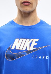 Κοντινή λήψη ενός ατόμου που φοράει μπλε μπλούζα Nike με μεγάλο λευκό και ριγέ λογότυπο Nike και τη λέξη "FRANCE" από κάτω.