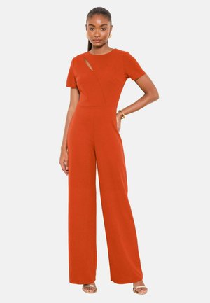 Kvinde iført en rustorange kortærmet jumpsuit med vide ben og en lille diagonal udskæring nær halsudskæringen, poserer med den ene hånd på hoften.