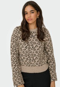 Beige sweater med et leopardmønster, ribbet rund hals og manchetter. Blød tekstur med en let kroppet pasform. Sorte bukser synlige.