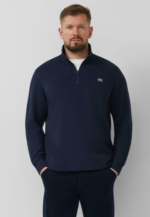 Mann mit Bart, der einen marineblauen Pullover mit Reißverschluss oben und dunkle Hosen trägt, steht mit den Händen in den Taschen vor einem grauen Hintergrund.