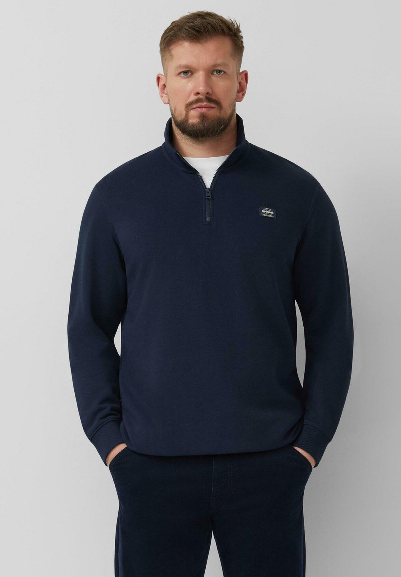 Mężczyzna z brodą, ubrany w granatowy sweter na zamek do połowy i ciemne spodnie, stoi z rękami w kieszeniach na szarym tle.