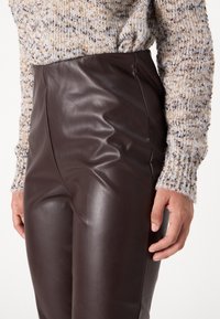 Pantalon taille haute en simili cuir marron avec une texture lisse, un design ajusté et des coutures verticales. Porté avec un pull texturé de couleur claire.