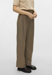 Pantaloni larghi marroni con una vestibilità su misura e una texture sottile, caratterizzati da pieghe frontali e una vita alta; abbinati a mocassini neri.