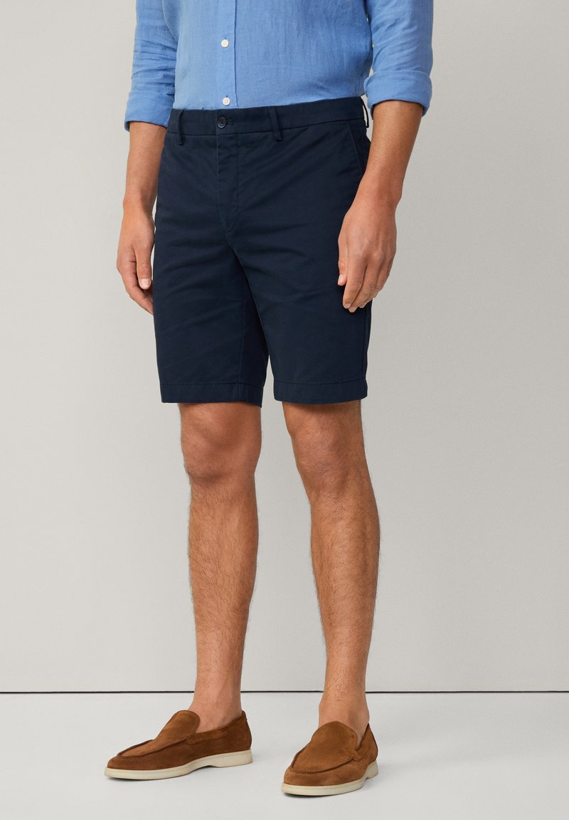 Shorts bleu marine dans un design ajusté, en coton, dotés de poches avant et d'une finition soignée à l'ourlet, associés à des chaussures à enfiler marron.