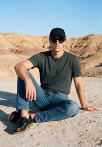 Homme portant une casquette noire, des lunettes de soleil, un t-shirt vert foncé et un jean bleu, assis jambes croisées sur un sol rocheux avec des collines désertiques en arrière-plan.