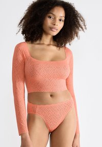 Kobieta z kręconymi włosami nosząca dopasowany crop top z długim rękawem oraz zestaw wysokich majtek w kolorze koralowym z delikatnym, teksturowanym wzorem.