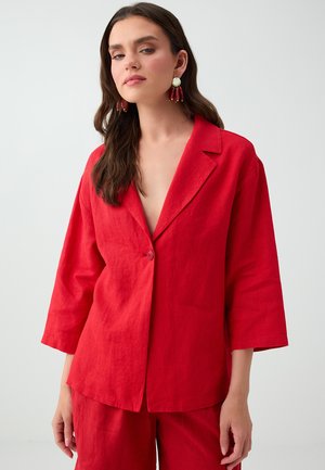 Frau mit langen braunen Haaren, die einen roten, geknöpften Blazer und passende Hose trägt, mit roten und goldenen hängenden Ohrringen vor einem einfarbigen Hintergrund.