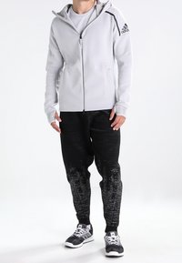 Helle graue Jacke mit Reißverschluss und Kapuze, dunkel gemusterte Jogginghose und schwarze Sportschuhe. Verfügt über das Adidas-Logo auf der Schulter.