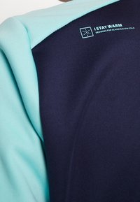 Chaqueta de forro polar con un diseño dividido en azul marino y turquesa claro. El texto en la parte posterior dice "ME MANTENGO CÁLIDO" con instrucciones de cuidado.