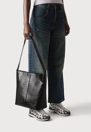 Sac en toile noir gaufré crocodile avec une longue bandoulière, porté sous le bras. Associé à un jean bleu lâche et des chaussures de sport à motif.