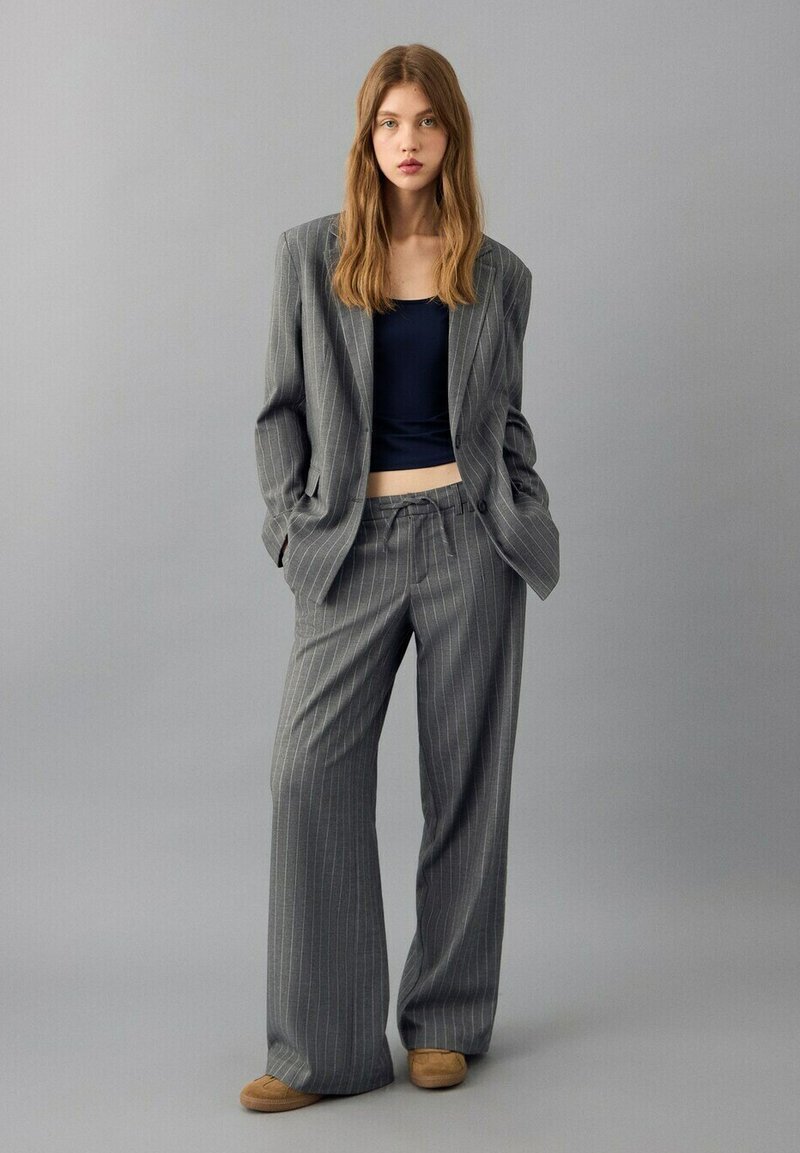 Jeune femme debout, les mains dans les poches, portant un costume ample gris à fines rayures, un haut noir court et des chaussures marron, sur un fond gris.