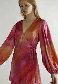Sukienka typu wrap z bufiastymi rękawami, wykonana z różowo-pomarańczowego materiału w stylu tie-dye, z detalem w postaci wiązania z boku oraz głębokim dekoltem w serek.