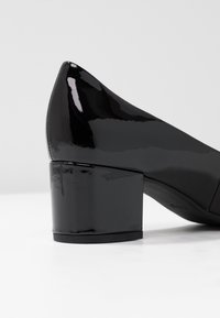 Talons en cuir verni noir, avec un bout pointu et un talon épais. Texture lisse et brillante avec un minimum de détails.