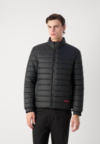 Zwarte gewatteerde jas met een hoge kraag, ritssluiting aan de voorkant, elastische manchetten en een rood logo-detail. Kenmerkt een horizontaal gestikt ontwerp en een gladde textuur.