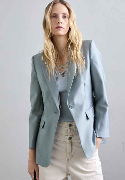 Blondynka z luźnymi falowanymi włosami, ubrana w jasnoniebieską marynarkę na prążkowanej bluzce i beżowych spodniach z wysokim stanem, stojąca na jednolitym szarym tle.