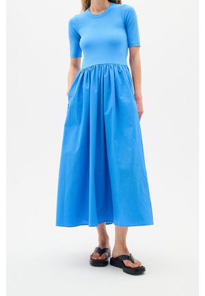 Donna che indossa un vestito aderente blu a maniche corte con gonna raccolta a mezza lunghezza e sandali platform neri, in piedi su sfondo bianco.
