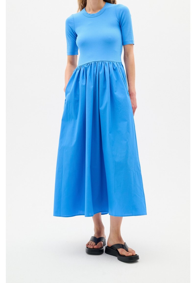 Femme portant une robe bleue ajustée à manches courtes avec une jupe mi-longue froncée et des sandales à plateforme noires, debout sur un fond blanc.