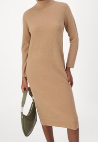 Robe marron en maille à col roulé avec manches longues et fentes latérales, associée à un sac à main vert. Côtelé texturé sur toute la surface.