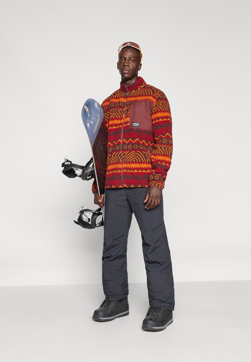 Red and orange patterned fleece jacket with a chest pocket, paired with black insulated pants and snow boots, holding a snowboard with bindings.