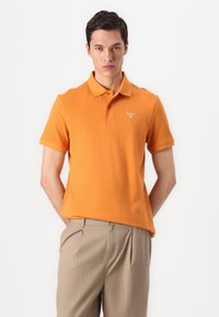 Πορτοκαλί polo μπλούζα από βαμβάκι, με γιακά και κοντά μανίκια. Συνδυάζεται με ανοιχτό μπεζ παντελόνι. Λογότυπο στο αριστερό στήθος.