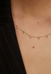 Collar de cadena de oro con piedras preciosas de varios colores (amarillo, verde, rosa) espaciadas uniformemente, utilizado sobre un fondo de tela oscura con rayas.