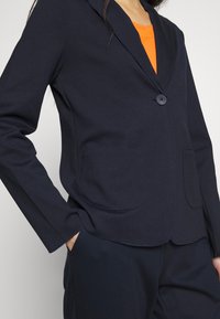 Donna che indossa un blazer blu navy con due tasche frontali su una maglietta arancione, con le mani nelle tasche dei pantaloni blu navy abbinati, tagliato all'altezza del collo e delle cosce.