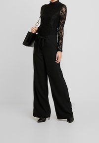 Svart spetsbodysuit med långa ärmar, i kombination med höga svarta byxor med vida ben och en frontslips. Håller en svart strukturerad väska.