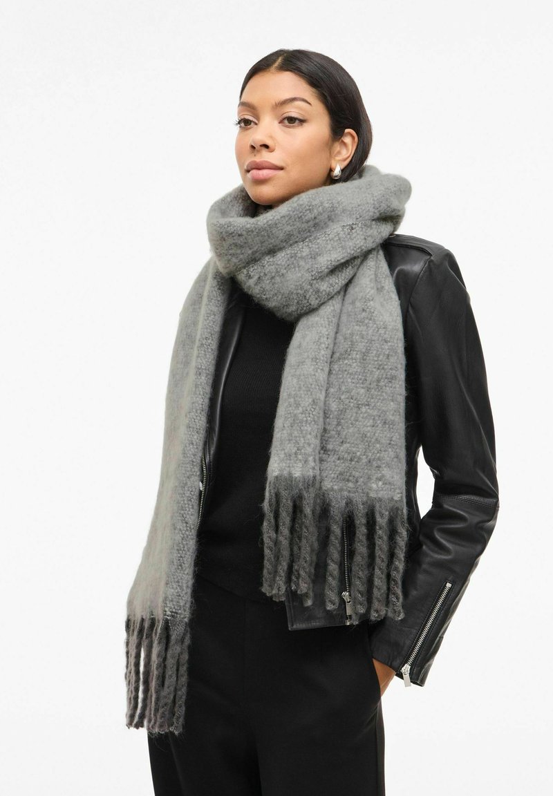Gråt uldtørklæde med en blød tekstur, der har frynser i kanterne og et hyggeligt fald, kontrasteret mod et sort outfit og en læderjakke.