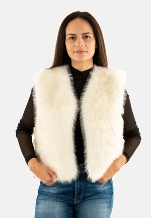 Donna con lunghi capelli castani che indossa un gilet bianco e morbido sopra una maglia nera a maniche lunghe e jeans blu, in piedi con le mani nelle tasche.