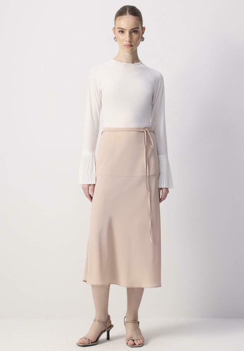 Γυναίκα που φοράει λευκό μπλουζάκι με μακριά μανίκια και πιετέ μανσέτες, μπεζ midi φούστα τύπου wrap και πέδιλα ανοιχτών δαχτύλων, στέκεται μπροστά σε απλό φωτεινό φόντο.