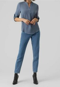 Blouse bleu clair à manches longues avec col en V et poignets retroussés, associée à un jean bleu ajusté et des bottines noires. Texture de tissu lisse.