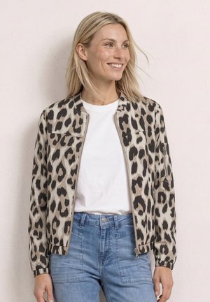 Femme souriante, portant une veste beige et noire à motif léopard sur une chemise blanche et un jean bleu clair, debout contre un mur pâle.
