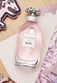 Clear glass perfume bottle with a rounded design, light pink liquid, silver cap, and a label reading "COACH DREAMS" featuring a carriage illustration.