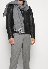 Mies, joka pitää mustaa nahkatakkia, harmaata kuvioitua huivia kaulassaan ja harmaita siistipö trousers, valkoista taustaa vasten.