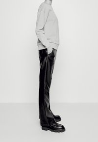 Persona che indossa pantaloni in pelle nera, scarpe nere chunky e una felpa a maniche lunghe di colore chiaro, in piedi di lato con le mani nelle tasche.
