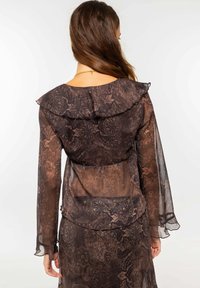 Blusa marrón transparente con mangas largas, escote con volantes y intrincado patrón de paisley. Diseño en capas con una textura suave y fluida.