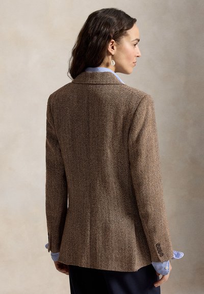 Woman with dark wavy hair wears brown herringbone blazer over light blue shirt, facing right against a neutral background.