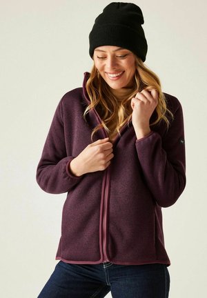 Uśmiechnięta kobieta w czarnej czapce typu beanie i fioletowej kurtce zapinanej na zamek, na beżowym golfie, trzymająca zamek blisko klatki piersiowej.