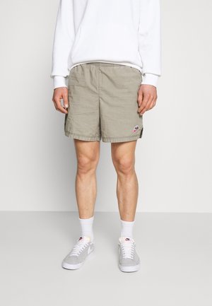Khakifarbene Shorts aus leichtem Stoff, mit lockerem Schnitt, Seitentaschen und einem kleinen Logopatch. Kombiniert mit weißen Socken und Turnschuhen.