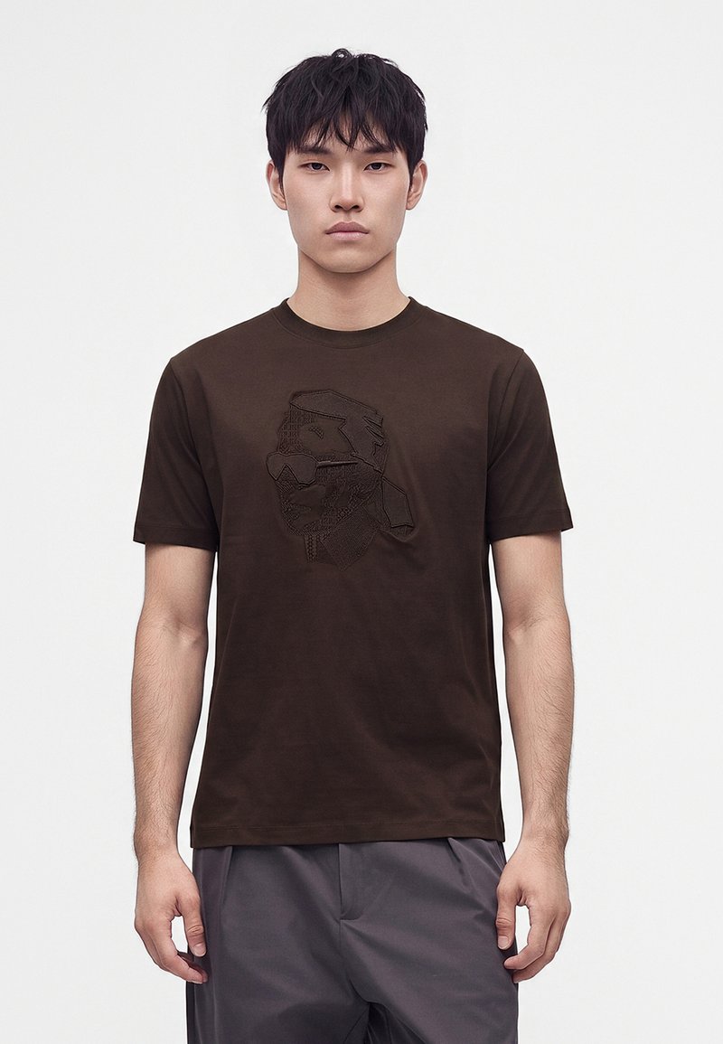 Νέος άνδρας που φοράει ένα σκουροκαφέ t-shirt με μια διακριτική κεντημένη εικόνα προσώπου μπροστά, στέκεται μπροστά σε ένα απλό λευκό φόντο.