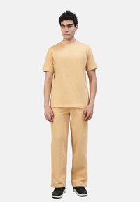 Beige Baumwoll-T-Shirt mit kurzen Ärmeln und Rundhalsausschnitt, kombiniert mit passenden lockeren Hosen, getragen mit schwarzen Sneakers.