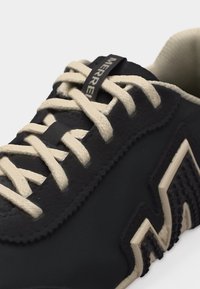 Baskets noires avec lacets et doublure beiges, arborant un logo "M" texturé beige et noir sur le côté ainsi que la marque "Merrell" sur la languette.
