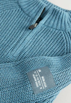 Lys blå strikket pullover med lynlås i nakken. Har struktureret syning og et stofmærke med produktinformation syet ind i siden.