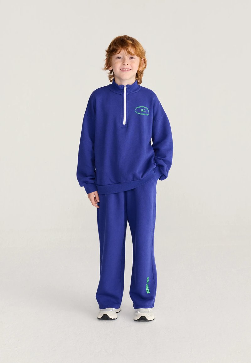 Enfant aux cheveux roux souriant, portant un survêtement bleu assorti et des baskets blanches sur un fond clair uni.
