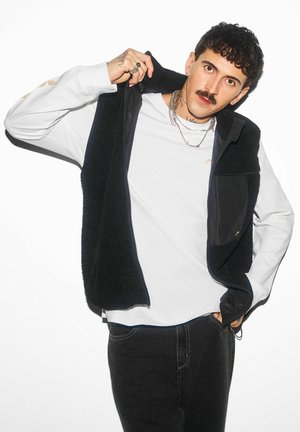 Mężczyzna z wąsami i tatuażami podnoszący czarną polarową kamizelkę, ubrany w białą koszulę z długim rękawem, czarne spodnie, srebrne naszyjniki i pierścionki na białym tle.