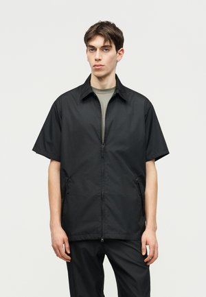 Jeune homme portant une veste noire à manches courtes avec fermeture éclair sur un t-shirt gris et un pantalon noir assorti, debout devant un fond uni.