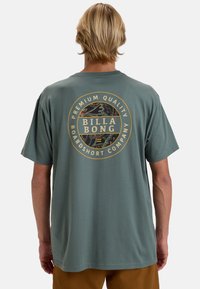 Grünes Baumwoll-T-Shirt mit einem kreisförmigen Logo in Gold und Schwarz auf dem Rücken, kurzem Ärmel und lässiger Passform.