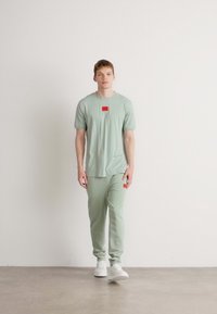 Camiseta de algodón verde claro y joggers a juego con un parche de logo rojo. El modelo lleva zapatillas blancas y está de pie frente a un fondo neutro.