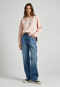 Felpa oversized rosa chiaro con logo argento, abbinata a jeans blu a vita alta e sneakers bianco e nero, in piedi su uno sfondo neutro.