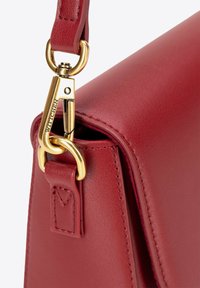 Hoek van een rode leren handtas met gestructureerde afwerking, gouden hardware met haaksluiting en stikseldetails. Merken zichtbaar op de hardware.